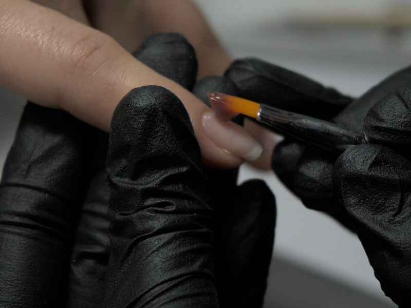 Black-gloved hands holding a fine brush against a fingernail