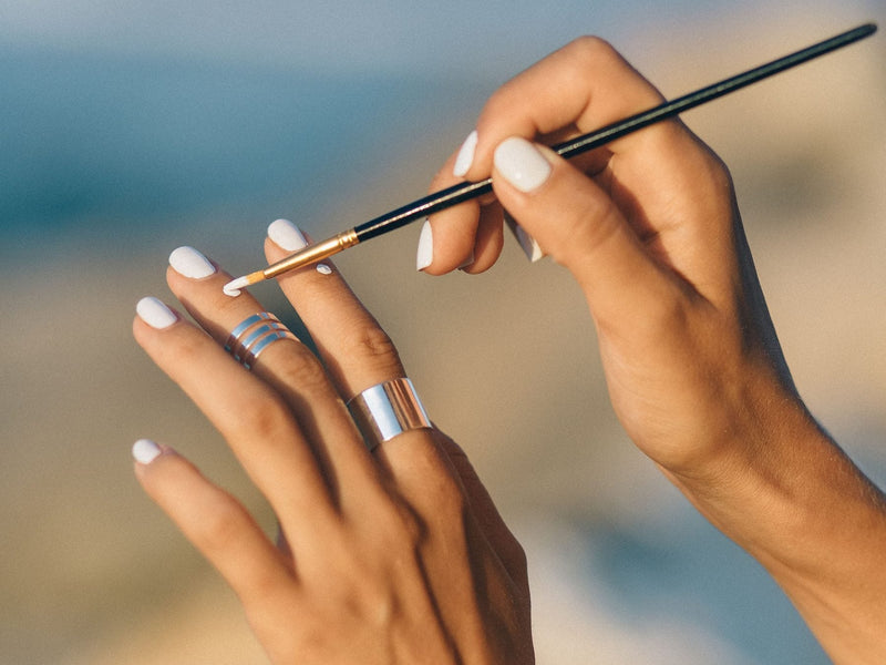 Two hands with white nails, one holding a thin nail art brush