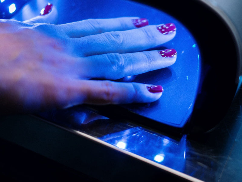 Hand with magenta nail polish curing inside a blue-lit nail lamp